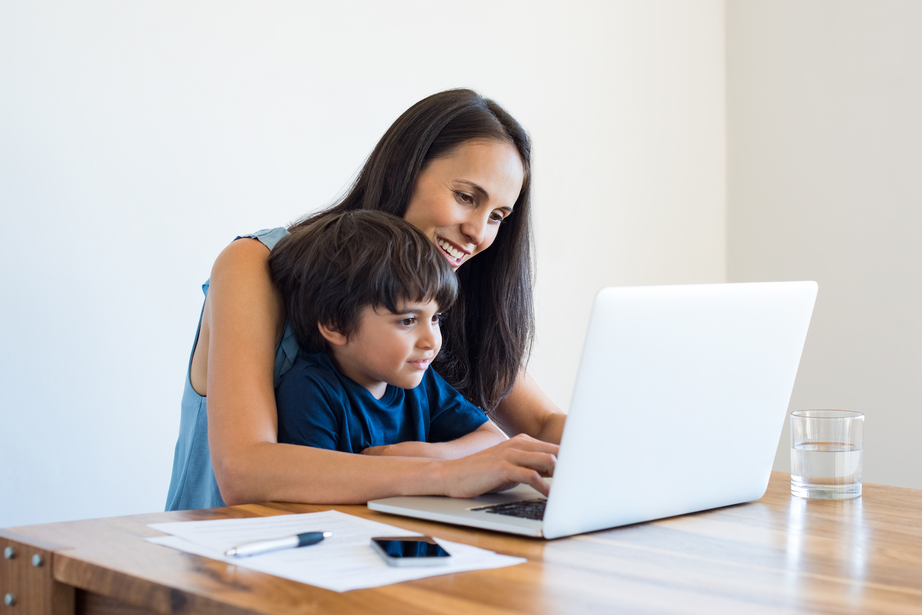 Contact Us Young mother and son sitting at table and using laptop at home. Smiling mom working at home with her child on the knees while writing an email. Young woman teaching little boy to use the computer.
